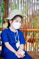 Kind mit weißer medizinischer Gesichtsmaske. Kind, das Huhn auf ihren Händen hält. Küken thront auf Schulter kleines Mädchen. ermöglicht es Kindern, sich dem Konzept der Nutztiere zu nähern.