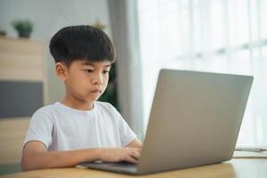 ein jung Junge ist Sitzung beim ein Tabelle mit ein Laptop im Vorderseite von ihn. er ist konzentriert auf das Bildschirm, möglicherweise Arbeiten auf ein Schule Zuordnung oder spielen ein Spiel foto