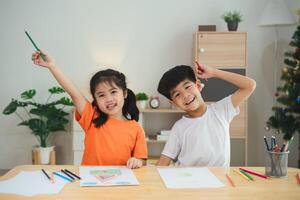 zwei Kinder sind Sitzung beim ein Tabelle mit Buntstifte und Zeichnung Papier. einer von das Kinder ist halten ein Bleistift und das andere ist halten ein Wachsmalstift. Sie sind beide lächelnd und scheinen zu Sein genießen sich foto