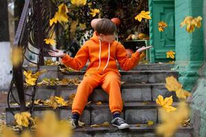 ein Junge im Orange Kleider sitzt auf das Schritte und sieht aus beim das Gelb fliegend Herbst Blätter. fallen hat kommen. foto