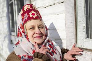 ein Alten Russisch Frau im ein traditionell Kopfschmuck, Kokoschnik, posiert gegen das Hintergrund von ein Dorf Haus. foto