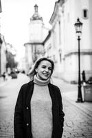 lächelnd Frau im schwarz Mantel und grau Sweatshirt Gehen Nieder Straße. ein heiter Frau, Balken mit ein warm Lächeln wie sie Spaziergänge Nieder das Straße, hervorrufen ein Sinn von Glück und Komfort. foto