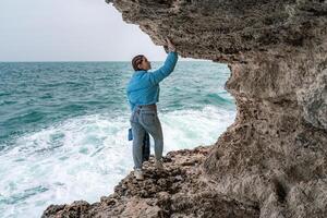 ein Frau im ein Blau Jacke steht auf ein Felsen über ein Cliff über das Meer und sieht aus beim das tobt Ozean. Mädchen Reisender ruht, denkt, Träume, genießt Natur. Frieden und Ruhe Landschaft, windig Wetter. foto