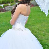 Nahansicht Porträt von jung schön Braut im ein Hochzeit Kleid Stehen auf ein Grün Feld und halten ein Weiß Regenschirm foto