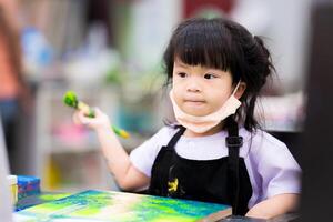 asiatisch wenig Mädchen ist Sitzung halten ein groß Pinsel und Gemälde Aquarell auf ein Leinwand, Kind genießt Lernen Kunst, süß Kind Freizeit Aktivität. Kinder alt 4 Jahre. foto