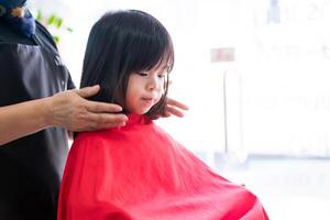 das Hände von ein Friseur sind Styling ein wenig asiatisch Mädchen Haar nach das Haarschnitt ist vollständig. das Friseur ist Überprüfung das Vollständigkeit von das arbeiten. Kind ist Sitzung und Schneiden ihr Haar beim Barbier foto