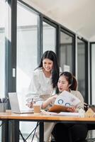 zwei Fachmann Frauen beschäftigt, verlobt im Überprüfung ein Geschäft Bericht zusammen im ein modern Büro Rahmen mit ein Laptop auf das Schreibtisch. foto