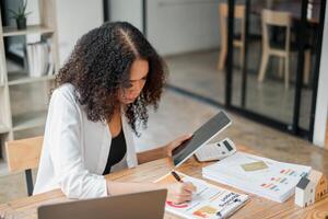 Fachmann Frau ist tief konzentriert auf finanziell Analyse, mit ein Taschenrechner und Tablette mit Diagramme und ein Laptop auf ihr Schreibtisch. foto