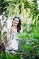 lächelnd zufrieden, ein Frau genießt ihr Kaffee brechen im ein üppig Garten, mit ihr Arbeit gelegt aus auf das Tabelle im Vorderseite von ihr. foto
