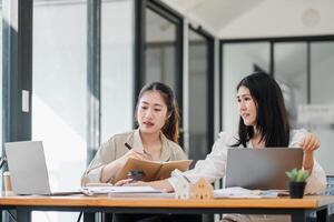 zwei Fachmann Frauen beschäftigt, verlobt im Diskussion, mit einer Schreiben im ein Notizbuch und das andere mit ein Laptop beim ein Büro Tisch. foto