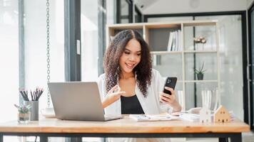 beschäftigt, verlobt Geschäftsfrau mit ein Smartphone im einer Hand während interagieren mit ein Laptop, umgeben durch Büro Lieferungen. foto