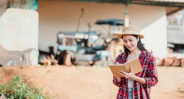 Farmer Frau im ein Stroh Hut und Plaid Hemd nimmt Anmerkungen auf ein Notizbuch mit ein Traktor im das Hintergrund. foto