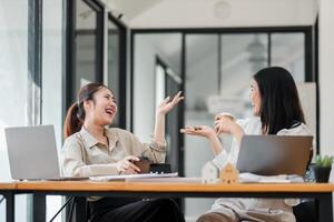 zwei freudig asiatisch Frauen engagieren im ein lebhaft Gespräch, einer gestikulieren animiert während Sitzung beim ein hölzern Tabelle mit Laptops im ein hell Büro. foto