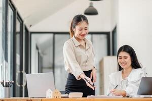 Stehen Frau lächelt beim ihr sitzend Kollege während diskutieren arbeiten, im ein modern Büro Rahmen mit Laptops und Arbeit Materialien auf das Schreibtisch. foto