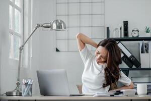 Frau nimmt ein entspannend strecken beim ihr Schreibtisch im ein gut beleuchtet, modern Zuhause Büro, nehmen ein brechen von ihr Arbeit auf das Laptop. foto