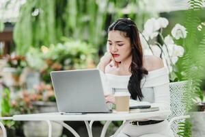 nachdenklich Frau ist tief konzentriert auf ihr Laptop Bildschirm, Arbeiten inmitten das üppig Flora von ein Gewächshaus Cafe. foto