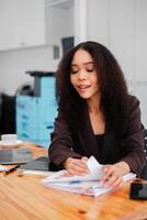 konzentriert afrikanisch amerikanisch Frau Sortierung durch Papierkram beim ihr Büro Schreibtisch, mit ein Laptop und Smartphone nahe. foto