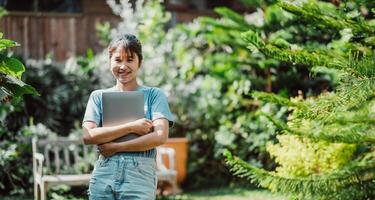 lächelnd Frau halten ein Laptop, Stehen im ein üppig Garten, symbolisieren Arbeitsleben Balance und Fernbedienung Arbeit Flexibilität. foto