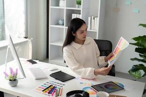 asiatisch Frau freiberuflich Grafik Designer Arbeiten mit Farbe Swatch Proben und Computer beim Schreibtisch im Zuhause Büro, jung Dame wählen Farbe Gamma zum Neu Design Projekt foto