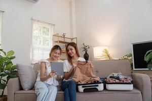 jung asiatisch Frau Verpackung Kleider zu das Koffer. Vorbereitung zum das Sommer- Urlaub. zwei Frauen sind Planung und mit mit Tablette zum Ausflug Buchung ein Ausflug und Portion zu bereiten Gepäck zu Reise foto