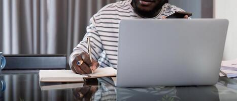 afrikanisch amerikanisch Mann Arbeiten mit Laptop Computer Fernbedienung während Sitzung beim Glas Tabelle im Leben Zimmer. schwarz Kerl tun freiberuflich während nehmen Anmerkungen Arbeit beim Zuhause Büro foto