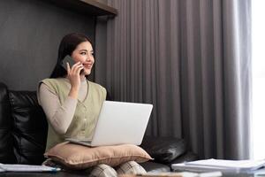 glücklich jung asiatisch freiberuflich Geschäft Frau Sitzung auf das Fußboden beim Zuhause Arbeiten auf Digital Tablette während Trinken Kaffee und sich unterhalten auf das Telefon foto