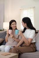 glücklich Senior Mutter mit Erwachsene Tochter Sitzung auf Couch und halten Tassen mit Kaffee oder Tee beim heim. genießen Familie Konzept foto