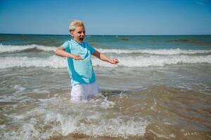 gut aussehend glücklich Teenager Junge von europäisch Aussehen mit blond Haar im Weiß kurze Hose, und ein Blau T-Shirt Sitzung und planschen im das Meer, und sieht aus zu das Kamera. Sommer- Familie Ferien konzept.sommer Reise konzept.kopie Raum. foto