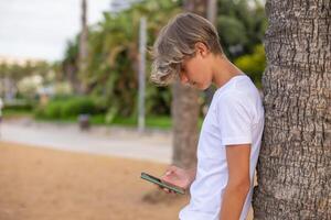 schließen oben gut aussehend Teenager Junge mit Handy, Mobiltelefon Telefon draußen im ein Park, Kopieren Raumseite Sicht. foto