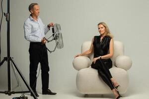 Studio Foto schießen. ein Frau posiert im ein Stuhl, und ein des Fotografen Assistent hält ein Studio Fan.