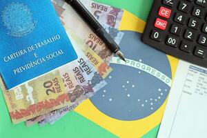 Brasilianer Arbeit Karte und Sozial Sicherheit Blau Buch und reais Geld Rechnungen mit Taschenrechner und Stift auf Flagge von föderativ Republik von Brasilien foto