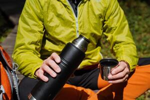 das Kerl hält im seine Hand ein Thermosflasche mit heiß Tee, Geschirr zum ein Wanderung, Mittagessen im Natur, ein thermo Glas mit Kaffee, Camping Ausrüstung Utensilien. foto