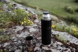 trinken Tee im Natur von ein Thermosflasche, öffnen Deckel, gießen Tee in ein Becher, Wanderung im das Berge, Camping im Natur, Tourist Teller, thermo Flasche. foto