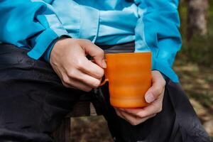 Damen Hände halt ein Becher von Kaffee im Natur, Essen auf ein Wanderung zu trinken heiß Tee von ein Tasse, ein Orange Glas zum Getränke, ein Plastik Becher Öko Produkt Pflege zum Natur. foto