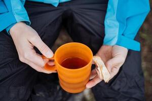 Frühstück Wandern Sandwich mit Käse im Hand halt ein Becher von Tee, trinken ein heiß trinken im Natur, warm oben mit warm Tee, köstlich Mittagessen beim das Campingplatz, Orange Glas foto