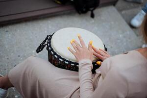 das Mädchen Theaterstücke das Trommel, Damen Hände schlagen das Rhythmus auf das Djemba, ein Straße Musiker im ein Kleid, ein Musical Instrument Schlagzeug, schön Nägel. foto