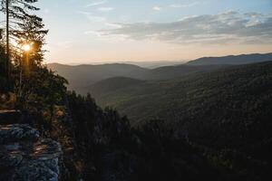 Aussicht von das Felsen Grat im das dunkel, Abend Beleuchtung, das Sonne Rollen gegenüber Sonnenuntergang, Blendung von Strahlen durch das Bäume, Berg Landschaft, Natur von Russland foto