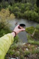 köstlich Tee mit Taiga Kräuter gegossen in ein Becher, ein Thermal- Glas mit ein heiß trinken, ein Hand hält ein Tasse von Tee gegen das Hintergrund von Natur, ein Ferien im das Berge foto