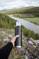 ein Thermosflasche von Stahl schwarz Farbe, das Ausrüstung von ein Tourist auf ein Wanderung, ein Vakuum Flasche behält Hitze. ein heiß trinken im das Wald. foto