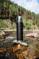 ein schwarz Metall Thermosflasche steht auf ein Stein im ein Berg Fluss, ein Vakuum Flasche zum heiß Getränke, Tee auf ein Wanderung im das Berge, ein thermo Flasche. foto