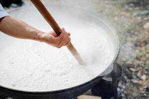 ein des Mannes Hand rührt sich Reis Haferbrei im ein groß Topf, ein Riese Portion von Essen, Kochen auf ein Lagerfeuer im das Wald, Morgen Haferbrei zum Frühstück. foto