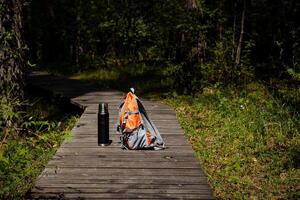 Camping Ausrüstung Lügen auf ein hölzern Pfad, ein Orange Rucksack, ein Metall Thermosflasche zum Tee, ein Wandern Tasche, Einzelheiten von ein Ausflug durch das Wald. foto