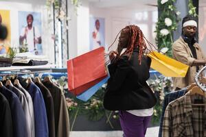 afrikanisch amerikanisch Frau Tanzen mit Freude während halten Einkaufen Taschen im Ausstellungsraum. glücklich jung Käufer Tragen mehrere Papier Pakete gefüllt mit ermäßigt Einkäufe im Kleidung Geschäft foto