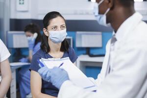 Nahansicht von ein Arzt im ein Labor Mantel und Gesicht Maske reden zu ein kaukasisch Frau im ein medizinisch Büro. Wirksam Kommunikation und personalisiert Gesundheitswesen sind offensichtlich im diese Klinik besuchen. foto