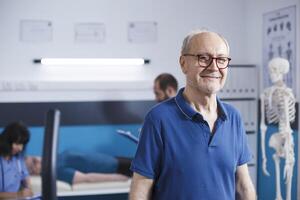 Alten Mann mit Brille Stehen im ein Physiotherapie Einrichtung. im Ruhestand Senior geduldig lächelnd beim Kamera wie er bekommt bereit zum seine Wiederherstellung beim Klinik zum Rehabilitation Behandlung durch Fitness. foto