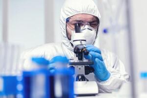 Mikrobiologe Forscher tragen ppe und Schutz Brille im Medizin Labor suchen durch Mikroskop. Wissenschaftler im schützend passen Sitzung beim Arbeitsplatz mit modern medizinisch Technologie während global Epidemie. foto