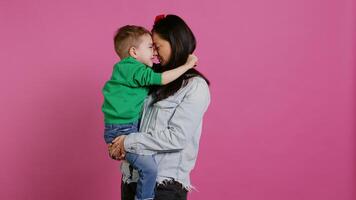 bezaubernd klein Junge umarmen seine Mutter und spielen um, haben Spaß gegen Rosa Hintergrund. jung Kleinkind posieren mit seine Mama halten ihn, Lachen und Sein heiter. Kamera b. foto