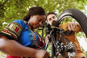 sportlich und eifrig schwarz Frau im Hof reparieren Fahrrad Umwerfer zum Sommer- Freizeit Radfahren. gesund engagiert sein afrikanisch amerikanisch weiblich Radfahrer unterstützt durch kaukasisch Mann im reparieren von Fahrrad Komponenten. foto