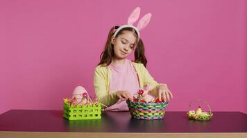 aufgeregt jung Mädchen arrangieren gemalt Eier im ein Korb zu bereiten zum Ostern Urlaub Feier, Erstellen festlich Anordnungen. spielerisch glücklich Kleinkind mit Hase Ohren, kreativ Aktivität. Kamera b. foto