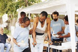 multiethnisch Nächstenliebe Gruppe Übergabe aus kostenlos Essen und Nahrung zu weniger glücklich und Behinderte. Porträt von schwarz Frau mit Blau T-Shirt assistieren das arm, Bedürftige afrikanisch amerikanisch Mann auf Krücken. foto